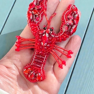 Peut inclure: Une broche homard rouge perlée, faite à la main. Le homard présente un travail de perles complexe, avec un corps, des pinces et des antennes détaillés. La broche est d'une teinte rouge vif, avec des accents de rouge plus clair et de perles transparentes. Mesure environ 10 cm de long.