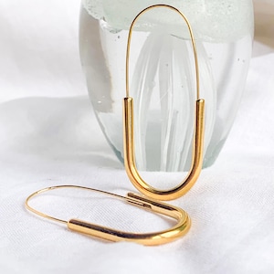 Puede incluir: Un par de pendientes de aro dorados en forma de U alargada. Los pendientes se exhiben sobre una superficie blanca, con un jarrón de cristal borroso en el fondo. Diseño sencillo y moderno.