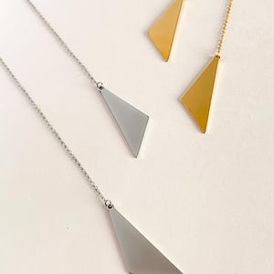 Puede incluir: Collares con colgantes geométricos en tonos plateados y dorados. Los collares presentan un colgante triangular suspendido de una delicada cadena. Los colgantes tienen una superficie lisa y plana. Las joyas se exhiben sobre un fondo claro.