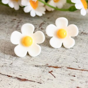 Könnte beinhalten: Zwei weiße Stecker-Ohrringe in Form von Gänseblümchen mit gelben Zentren. Die Ohrringe liegen auf einer verwitterten, hellen Holzoberfläche, mit einem verschwommenen Hintergrund aus Gänseblümchenblüten.