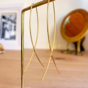 Peut inclure: Une paire de boucles d'oreilles dorées, allongées et incurvées, suspendues à un présentoir doré. Les boucles d'oreilles ont un design minimaliste, avec une apparence fine et lisse. Le support est rectangulaire à la base et possède une barre horizontale pour les boucles d'oreilles.