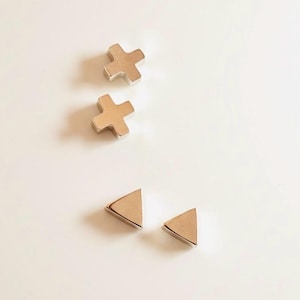 Peut inclure: Ensemble de quatre boucles d'oreilles dorées. Deux sont en forme de croix et deux sont triangulaires. Les boucles d'oreilles sont présentées sur une surface blanche. Elles sont petites et semblent être en métal.