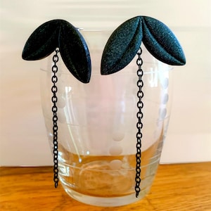 Op de afbeelding: Een paar donkergroene oorbellen in de vorm van bladeren met een getextureerde afwerking. Elke oorbel heeft een ketting die aan het bladontwerp hangt. De oorbellen worden getoond voor een helder glas.