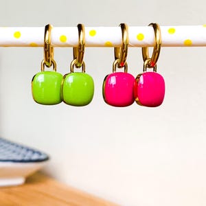 Op de afbeelding: Twee paar gouden oorringen met hangende vierkante bedels. Het ene paar heeft groene bedels en het andere paar heeft roze bedels.