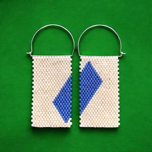 Puede incluir: Un par de pendientes hechos a mano con un diseño geométrico. Cada pendiente tiene forma rectangular con fondo crema y un patrón de rombos azules. Cuelgan de aros plateados.