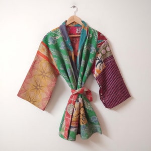 May include: A patchwork kimono robe with a tie closure, featuring a variety of patterns and colours. The robe includes green, red, yellow, and purple. The wide sleeves and the robe are hanging on a wooden hanger.