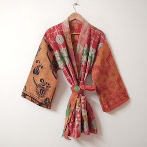 May include: A vibrant, patterned kimono robe in red, orange, and green, suspended on a wooden hanger. The robe has a tie belt and detailed designs on the sleeves and body. The background is a plain white wall.