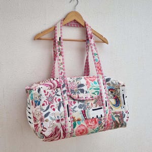 May include: A floral patchwork duffel bag in pink, red, and green hues. The bag, with a rectangular shape, features a small front pocket and pink striped handles. It is suspended from a wooden hanger. The bag's dimensions are approximately 40 cm in length.