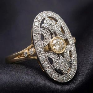 May include: An oval-shaped antique ring with a central, round, light-colored diamond. The ring features a gold band and a silver-colored filigree design encrusted with numerous small diamonds. The ring is set against a dark background.