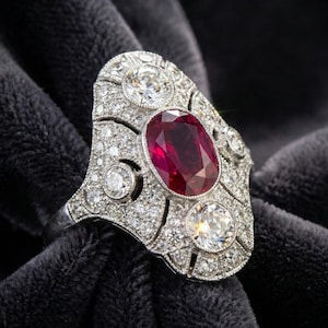May include: An ornate, vintage-style ring featuring a large, oval-cut ruby center stone. The ring is encrusted with numerous small, round diamonds, set in a white metal, likely platinum or white gold. The ring is displayed against a dark, velvety background.