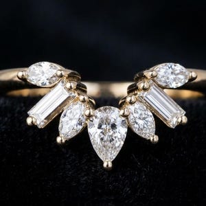 May include: A gold ring with a curved design, featuring a central pear-shaped diamond and surrounding baguette and marquise-cut diamonds. The ring is set against a dark background, highlighting the sparkle of the gemstones.