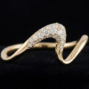 May include: A gold ring with a flowing, wave-like shape. The crest of the wave is set with numerous small diamonds. The ring's design is accentuated by the dark, black background.