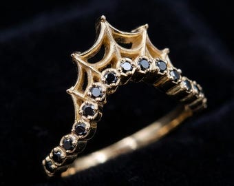 Puede incluir: Un anillo de oro con una serie de pequeñas piedras redondas de color negro engastadas a lo largo de una banda en forma de V. La parte superior del anillo muestra una estructura metálica elevada, puntiaguda y similar a una telaraña.