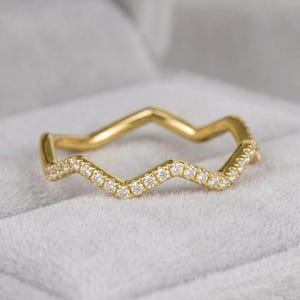 Peut inclure: Une bague dorée avec un motif en zigzag. La bague est ornée de petits diamants transparents le long du motif en zigzag. La bague est posée sur un fond gris doux.