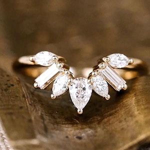 May include: A gold ring with a unique design featuring multiple clear, sparkling gemstones. The ring has a central pear-shaped stone, surrounded by smaller, marquise and baguette-cut stones, creating a fan-like arrangement. The band is a simple, polished gold.