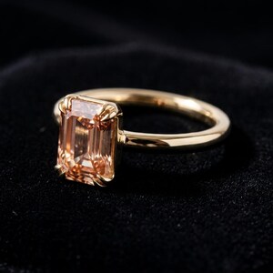 May include: A gold ring featuring a rectangular emerald-cut peach-colored gemstone set in a four-prong setting. The band is smooth and polished, showcasing a warm metallic tone that complements the light orange hue of the central stone.
