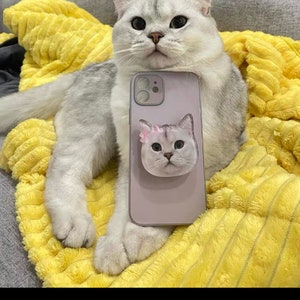 May include: A silver and white cat lies on a yellow blanket, beside a lilac phone case featuring a cat face design. The phone case has a pop-up grip with a cat face and a pink flower.
