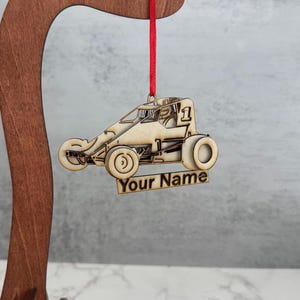 May include: A wooden ornament shaped like a race car, with the number 1 on top. The ornament is light brown and has the text "Your Name" on the bottom. It hangs from a red ribbon.