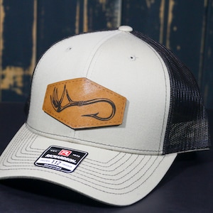 May include: A beige and black trucker hat with a brown leather patch featuring a deer antler and a fishing hook. The hat is made by Richardson 112.