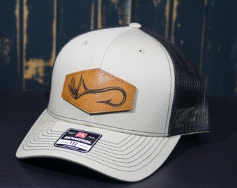 Op de afbeelding: Een beige en zwarte trucker cap met een bruine leren patch met een hertengewei en een vishaak. De pet is gemaakt door Richardson 112.