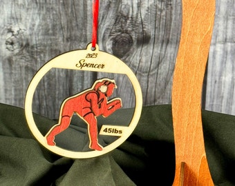 May include: A wooden Christmas ornament with a red and black illustration of a wrestler in a crouching position. The ornament is inscribed with the text "2023 Spencer 45lbs".