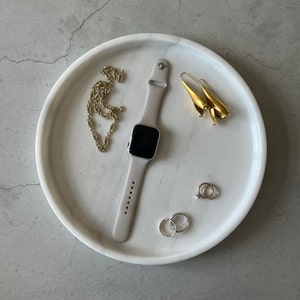 Pode incluir: Uma bandeja de m&aacute;rmore branco com um colar de corrente de ouro, um par de brincos de ouro, um anel de prata, um anel de ouro e um smartwatch preto e prateado.