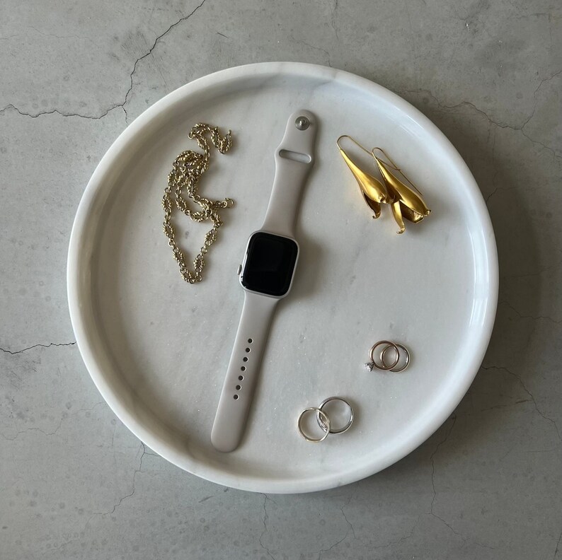 Pode incluir: Uma bandeja de m&aacute;rmore branco com um colar de corrente de ouro, um par de brincos de ouro, um anel de prata, um anel de ouro e um smartwatch preto e prateado.