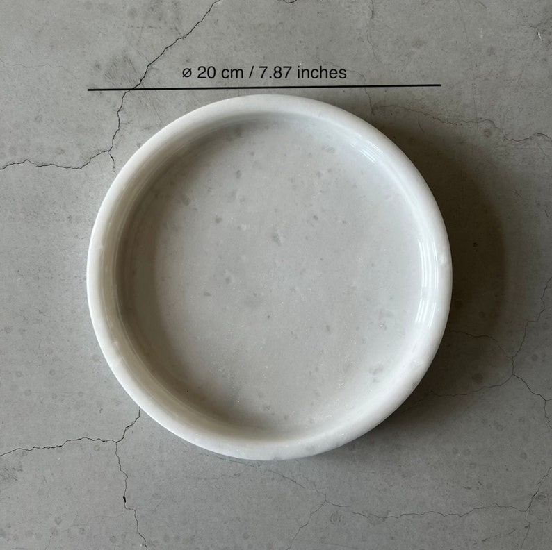 Pode incluir: Bandeja redonda de m&aacute;rmore branco com um di&acirc;metro de 20 cm. A bandeja &eacute; lisa e tem uma superf&iacute;cie ligeiramente texturizada.