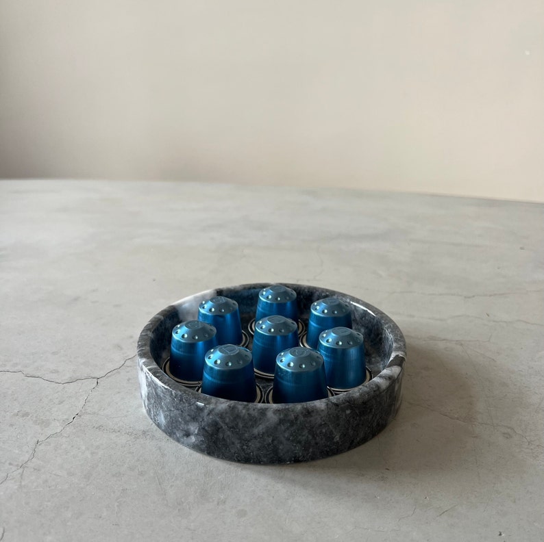 May include: A round, gray marble dish with a blue and white design holds nine blue coffee pods.