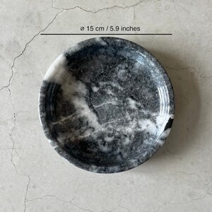 May include: A round, black and white marble bowl with a diameter of 5.9 inches. The bowl has a smooth, polished surface and a slightly raised rim.