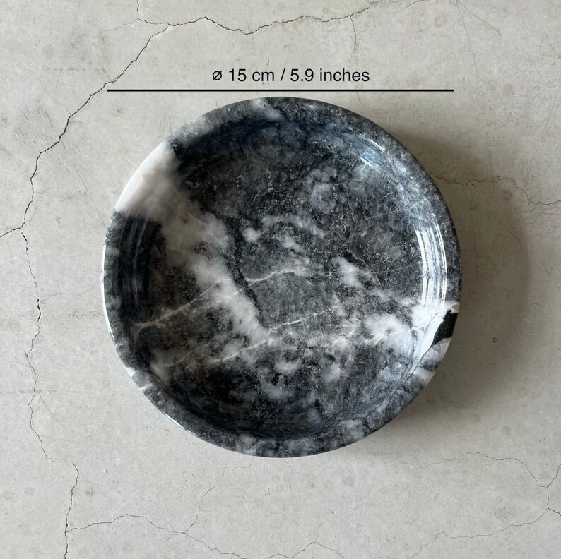 May include: A round, black and white marble bowl with a diameter of 5.9 inches. The bowl has a smooth, polished surface and a slightly raised rim.