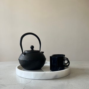Pode incluir: Uma bandeja de servir de m&aacute;rmore branco com uma chaleira de ferro fundido preto e duas x&iacute;caras de ch&aacute; de cer&acirc;mica preta.