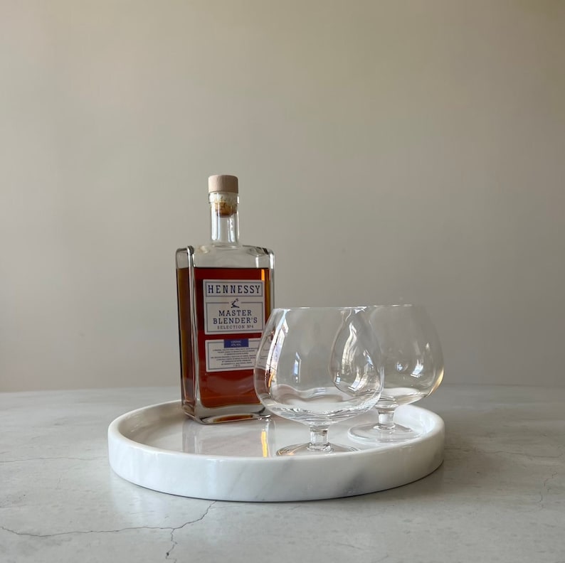 Pode incluir: Uma bandeja redonda de m&aacute;rmore branco com dois copos de conhaque de vidro transparente e uma garrafa de Hennessy Master Blender's Cognac.