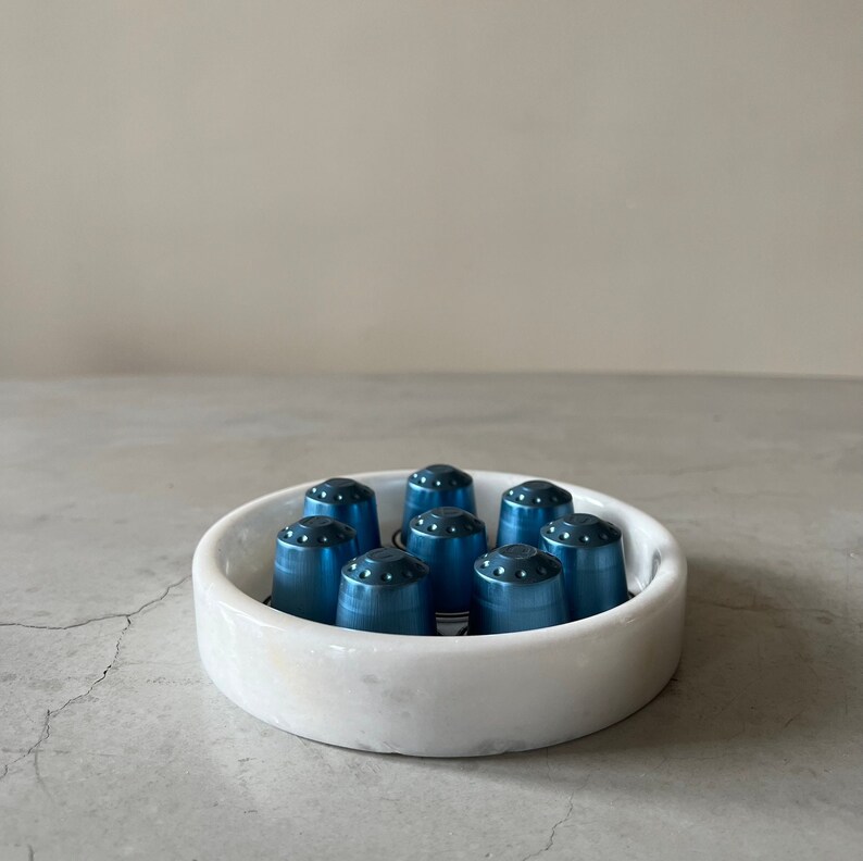 Pode incluir: Um prato de m&aacute;rmore branco com um design azul e branco cont&eacute;m dez c&aacute;psulas de caf&eacute; de metal azul.