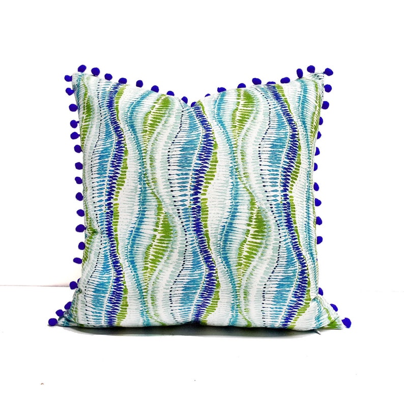Peut inclure: Un coussin d&eacute;coratif avec un motif ondul&eacute; bleu, vert et blanc. Le coussin est bord&eacute; d'une garniture de pompons bleus.