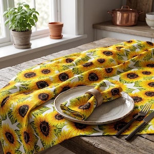May include: A table runner and matching napkin featuring a vibrant sunflower pattern with yellow petals, dark brown centers, and green leaves on a white background. The napkin is folded and held by a wooden ring on a ceramic plate with silverware.
