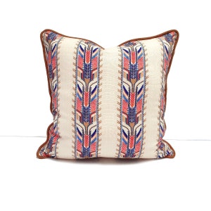 Peut inclure: Un coussin blanc avec une bordure en cuir marron et un motif géométrique dans les tons de rouge, bleu et beige.