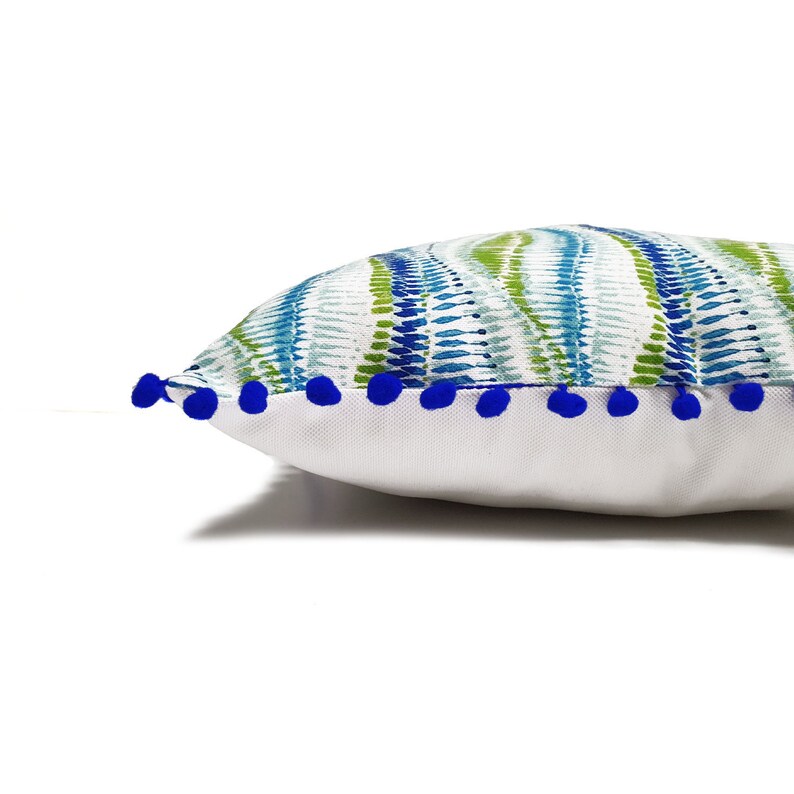 Peut inclure: Un oreiller blanc avec un motif abstrait bleu et vert et une bordure de pompons bleus.