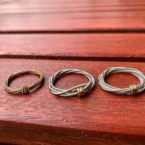 Könnte beinhalten: Drei Ringe aus Gitarrensaiten. Die Ringe sind silber und goldfarben. Die Ringe liegen auf einer Holzoberfläche.