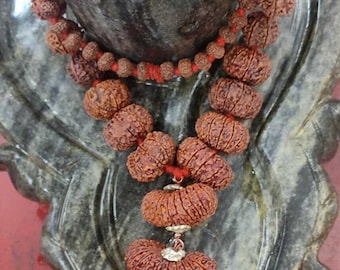 May include: A brown and red beaded necklace with a silver pendant. The necklace is made of small, round beads and is strung on a red cord. The pendant is a small, silver charm with a design on it. The necklace is lying on a gray, textured surface.