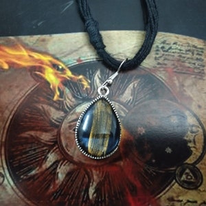 Może przedstawiać: Srebrny naszyjnik z wisiorem w kształcie łzy, ozdobiony czarnym i złotym kamieniem tygrysiego oka. Naszyjnik jest na czarnym sznurku i wisi nad starą książką z ilustracją smoka.