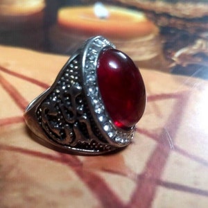 May include: A silver ring with a large, oval-shaped, red gemstone set in the center. The ring has intricate details and a band that is slightly wider than the stone.