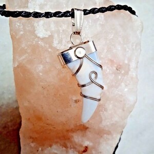 May include: A pendant necklace featuring a white, tooth-shaped stone wrapped in silver wire. The pendant hangs from a black cord. The stone has a silver cap and is set against a light pink background.