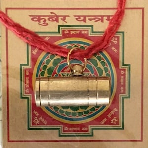 May include: A gold-coloured metal cylinder pendant hangs from a red string. The pendant is surrounded by a colourful mandala with Sanskrit text. The text reads "Kubera Yantra" and includes the phrase "Om Indray Namah".