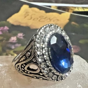 May include: Silver-toned ring featuring a large, faceted, oval-shaped blue gemstone surrounded by smaller clear stones. The ring has an ornate, detailed design on the band. The ring is set on a white surface.
