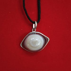 May include: A silver pendant with a white agate stone set in a diamond-shaped frame. The pendant is hanging from a black cord.