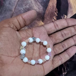 May include: White beaded bracelet with interspersed colorful beads. The bracelet features large, round white beads and smaller, translucent beads in shades of blue, pink, and yellow. A delicate piece of jewelry.