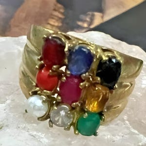 Op de afbeelding: Een gouden ring met een cluster van negen edelstenen. De stenen zijn rood, oranje, geel, groen, blauw, zwart, wit en twee tinten roze.