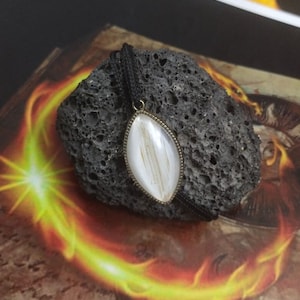 May include: A white oval-shaped pendant with a black cord. The pendant has a silver-colored metal frame and a white stone with a striated pattern.