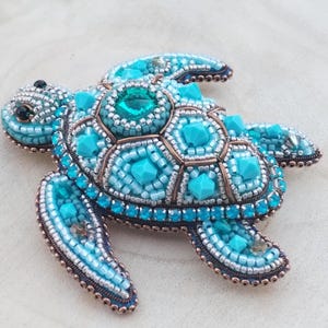 May include: A sea turtle brooch crafted with intricate beadwork in shades of turquoise, light blue, and silver. The shell features a central faceted teal crystal surrounded by geometric turquoise beads and metallic copper-toned wire accents.